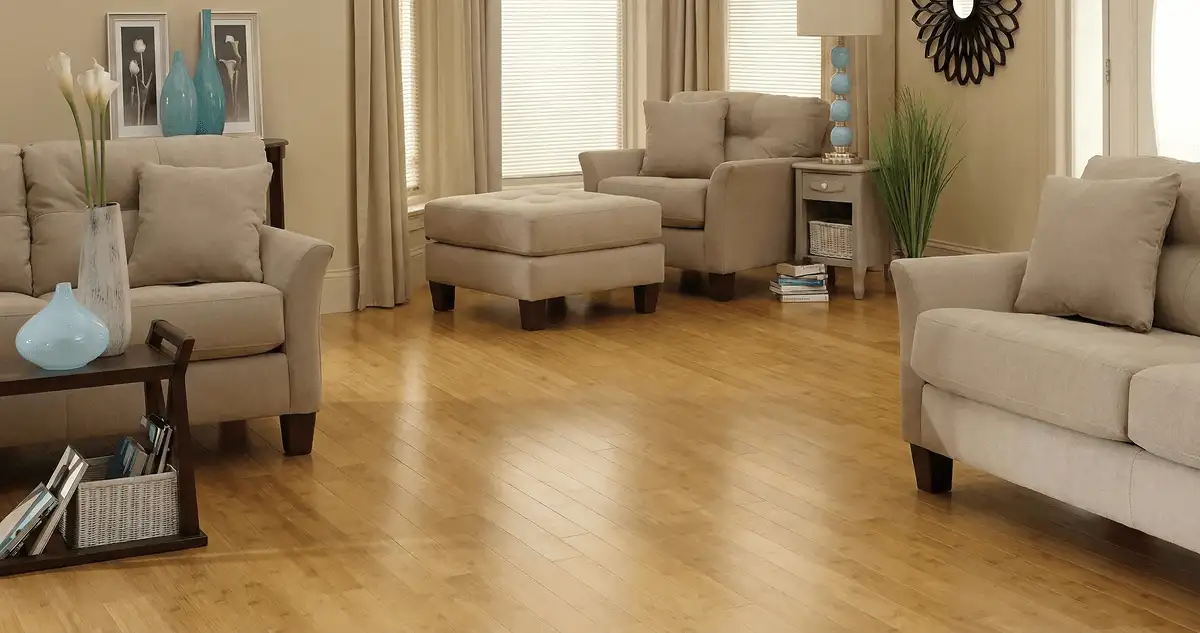 A living room with professionally installed bamboo flooring, beige sofas, a matching ottoman, a side table with a blue lamp, vases, framed pictures, and large windows with blinds and curtains in Oakland, CA