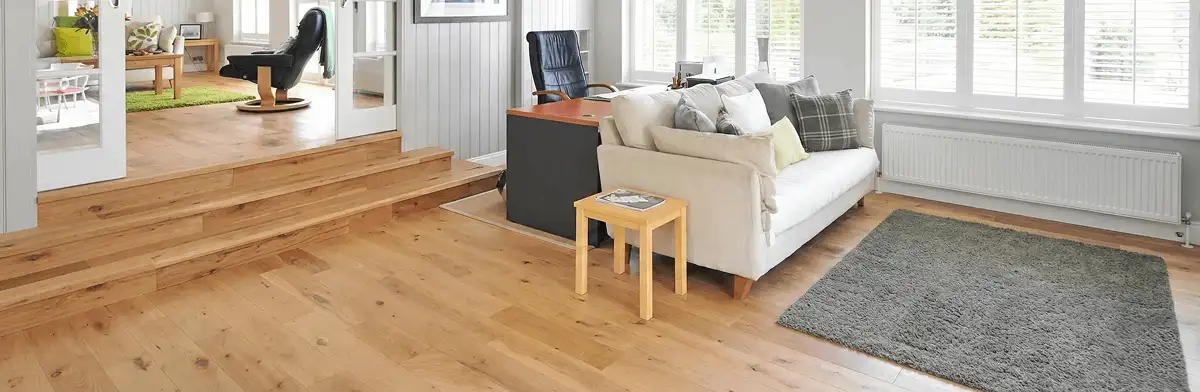 A modern living room with professionally installed laminate flooring, a beige sofa, a small side table, a gray rug, and large windows with white shutters in Oakland, CA